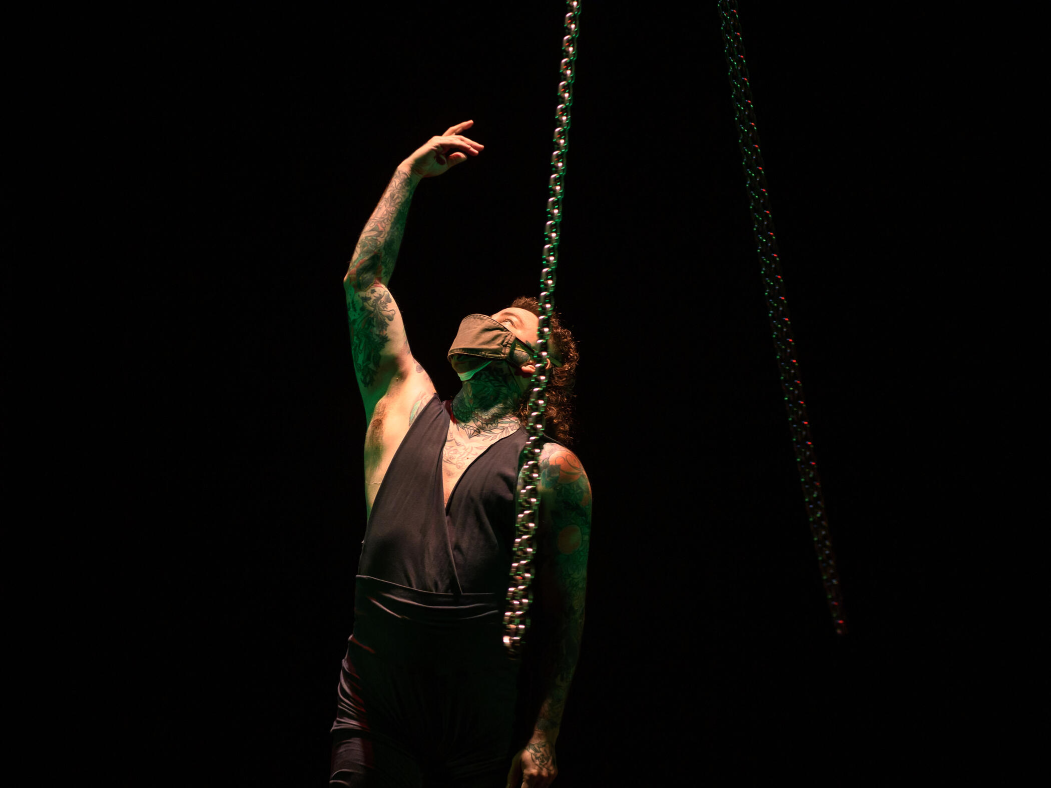 A performance photo of Alec standing between circling chains, gesturing with one arm up and looking up.