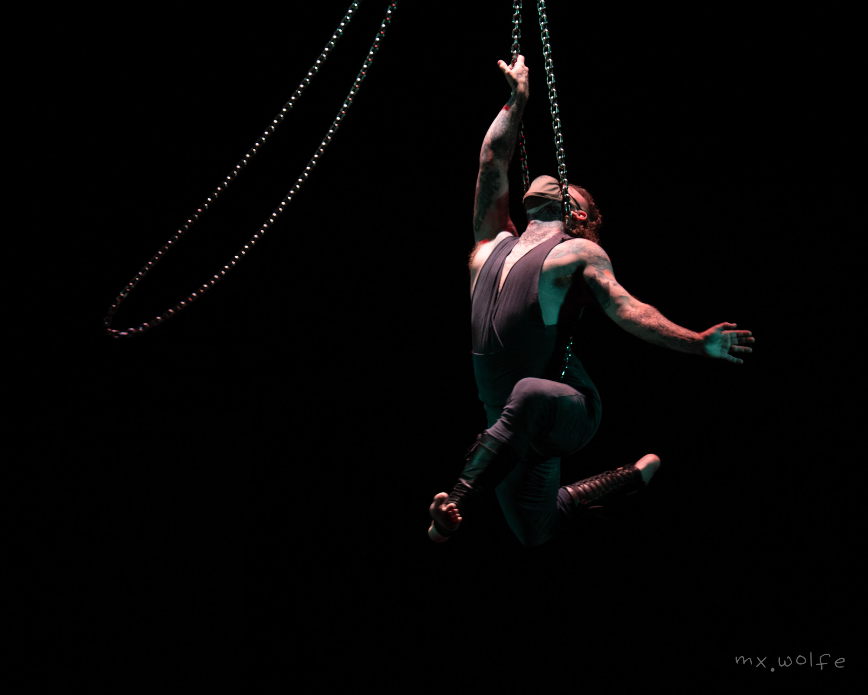 Alec suspended upright by one hip on one chain, his neck locked between the two sides with one arm open, one balancing above his head, the other chain circling him.